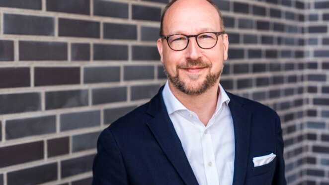 Lächelnder Mann mit Brille und dunklem Jackett steht vor einer Backsteinmauer mit HANSETRANS-Logo.