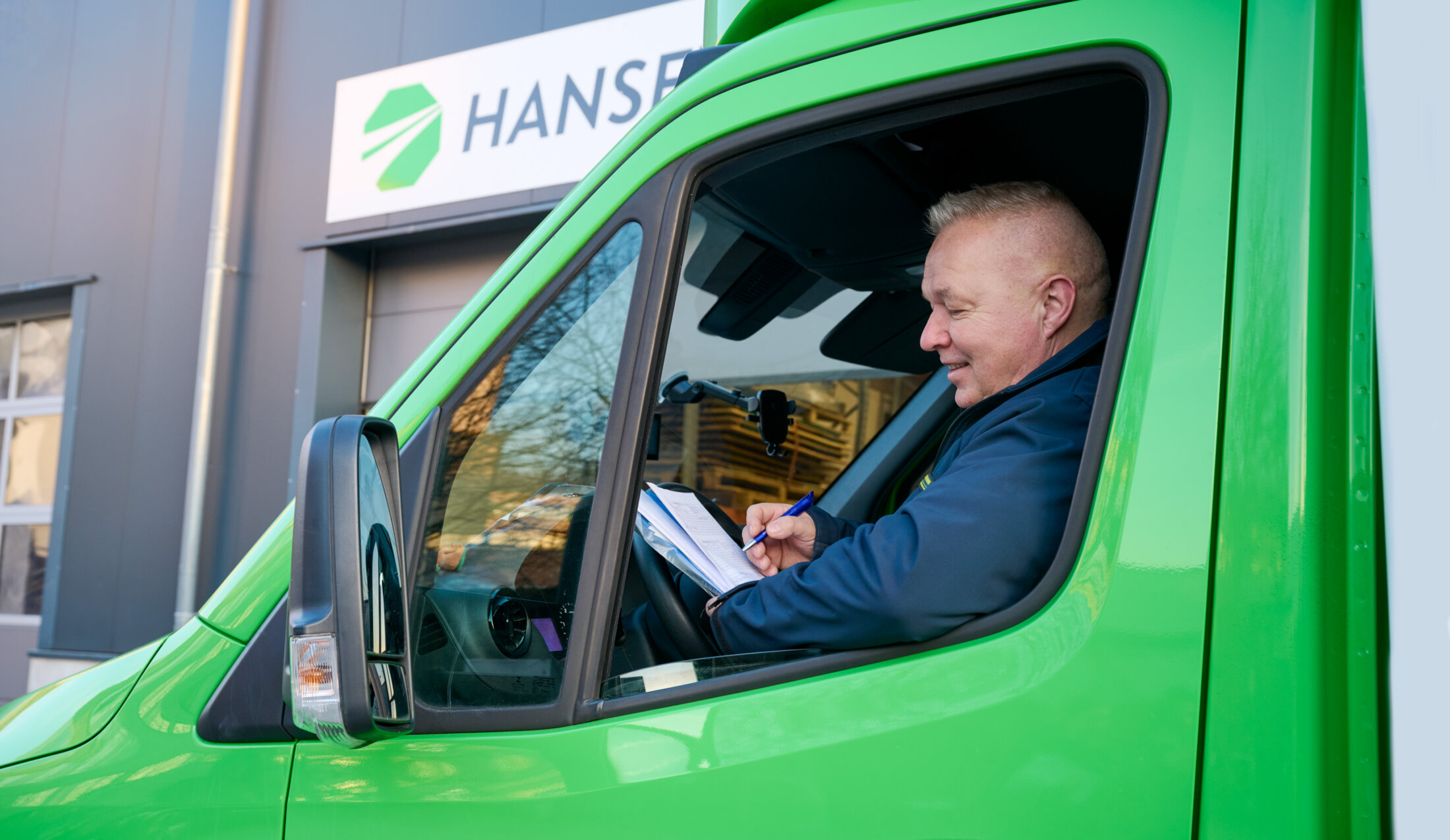 Ein Mann sitzt lächelnd in einem grünen LKW-Führerhaus und schreibt auf einem Klemmbrett vor einem Gebäude mit der Aufschrift „HANSETRANS“ – bereit für professionelle Entrümpelungs- und Haushaltsauflösungsdienste.