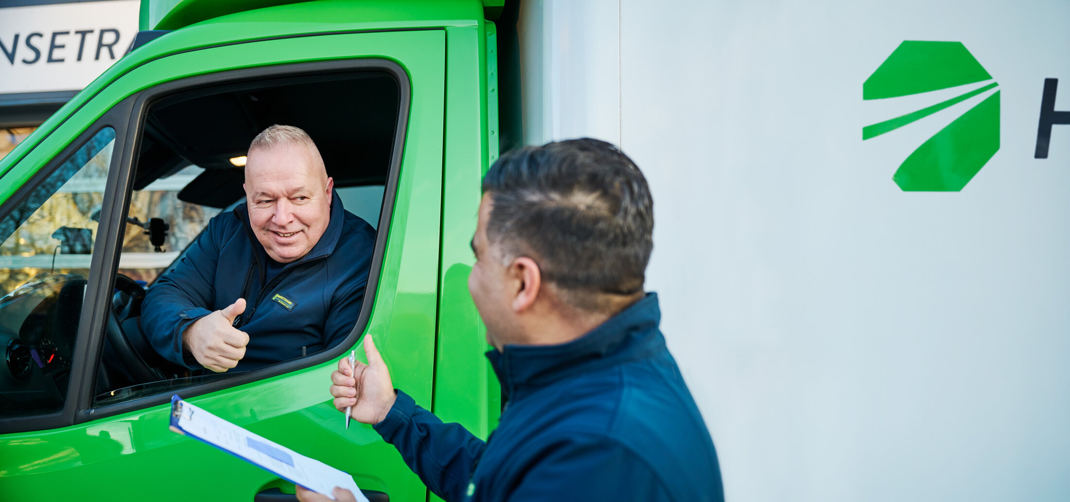 Ein Lkw-Fahrer von HANSETRANS zeigt einem Kollegen, der neben einem grünen Lieferwagen ein Klemmbrett hält, den Daumen nach oben.