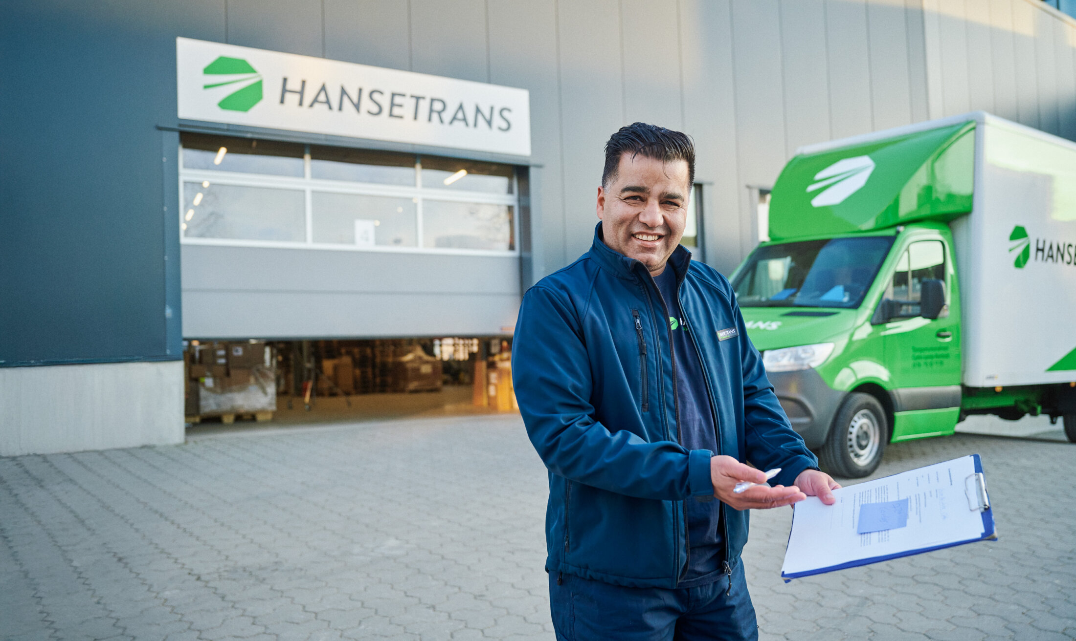 Lächelnder Zusteller mit Klemmbrett vor einem HANSETRANS-Lager und einem Lieferwagen mit Markenlogo, bereit, Ihnen bei Ihrem Privatumzug zu helfen.