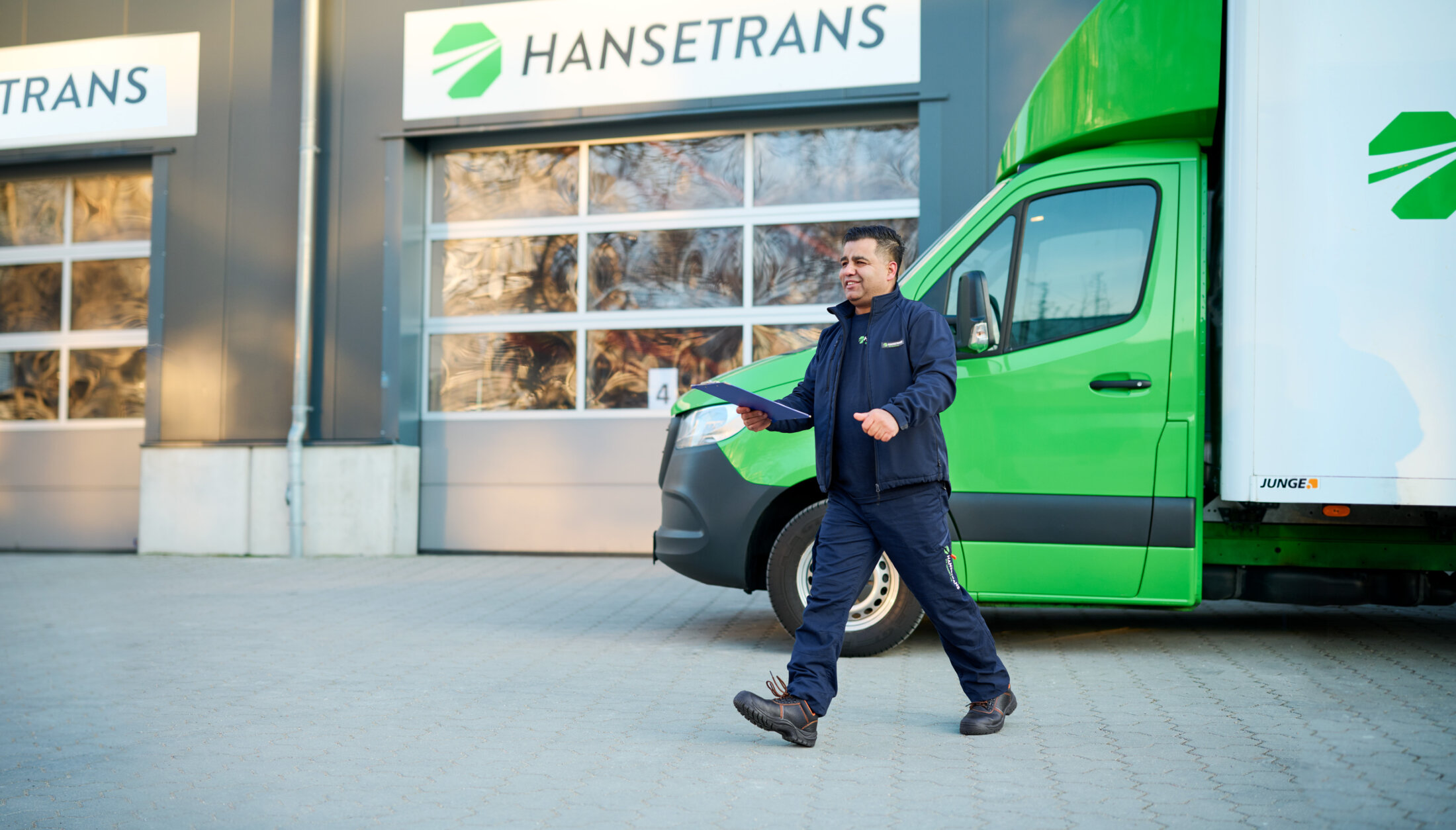 Ein Zusteller geht mit einem Klemmbrett neben einem grünen LKW von HANSETRANS, dem Vertreter des vertrauenswürdigen Transportdienstleisters.
