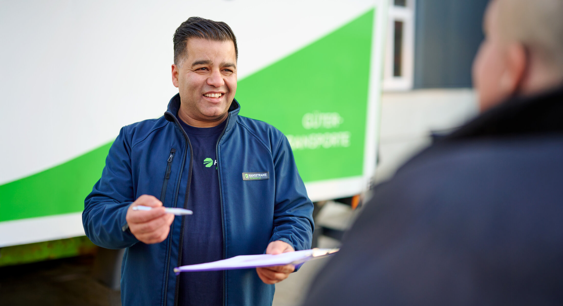 Lächelnder Lieferarbeiter mit Klemmbrett und Stift steht neben einem grün-weißen HANSETRANS-LKW und spricht mit einer Person über professionelle Entrümpelungsdienste.