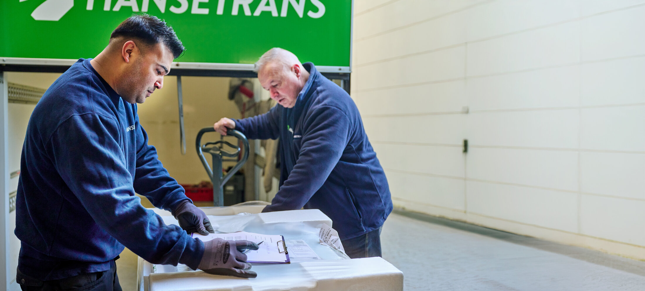 Zwei Männer in Arbeitsuniformen überprüfen ein Klemmbrett und schieben Pakete vor einen LKW von HANSETRANS, der einen professionellen Möbeltransportservice bietet.