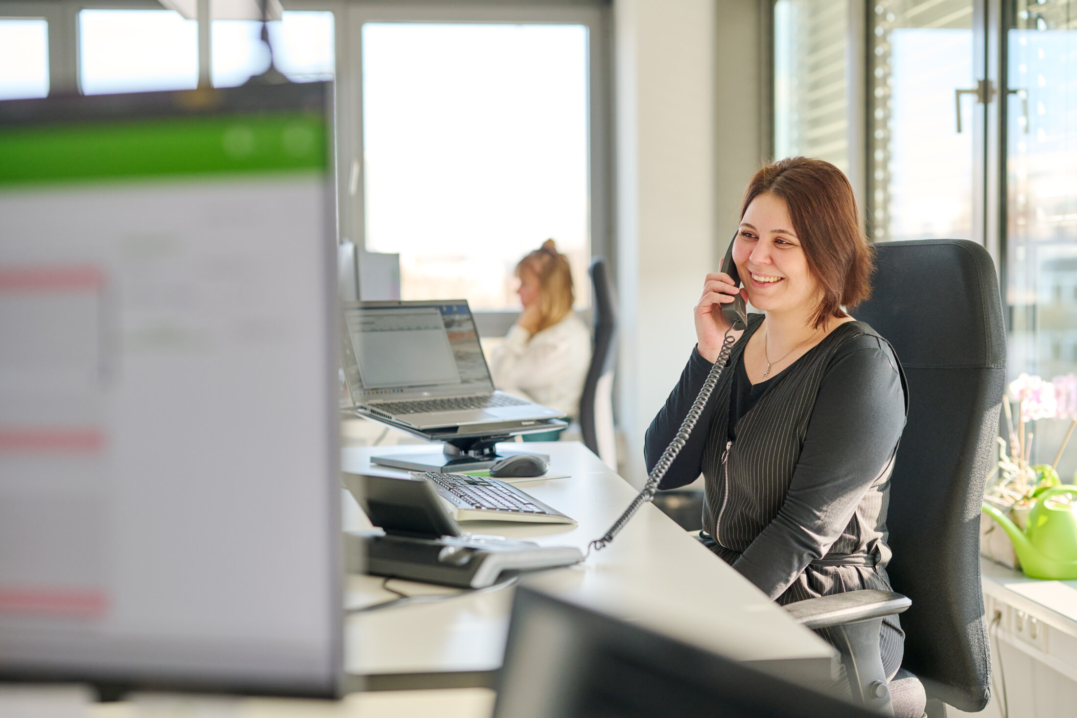 Frau sitzt an einem Schreibtisch in einem Büro bei HANSETRANS und lächelt, während sie mit einem schnurgebundenen Telefon telefoniert.
