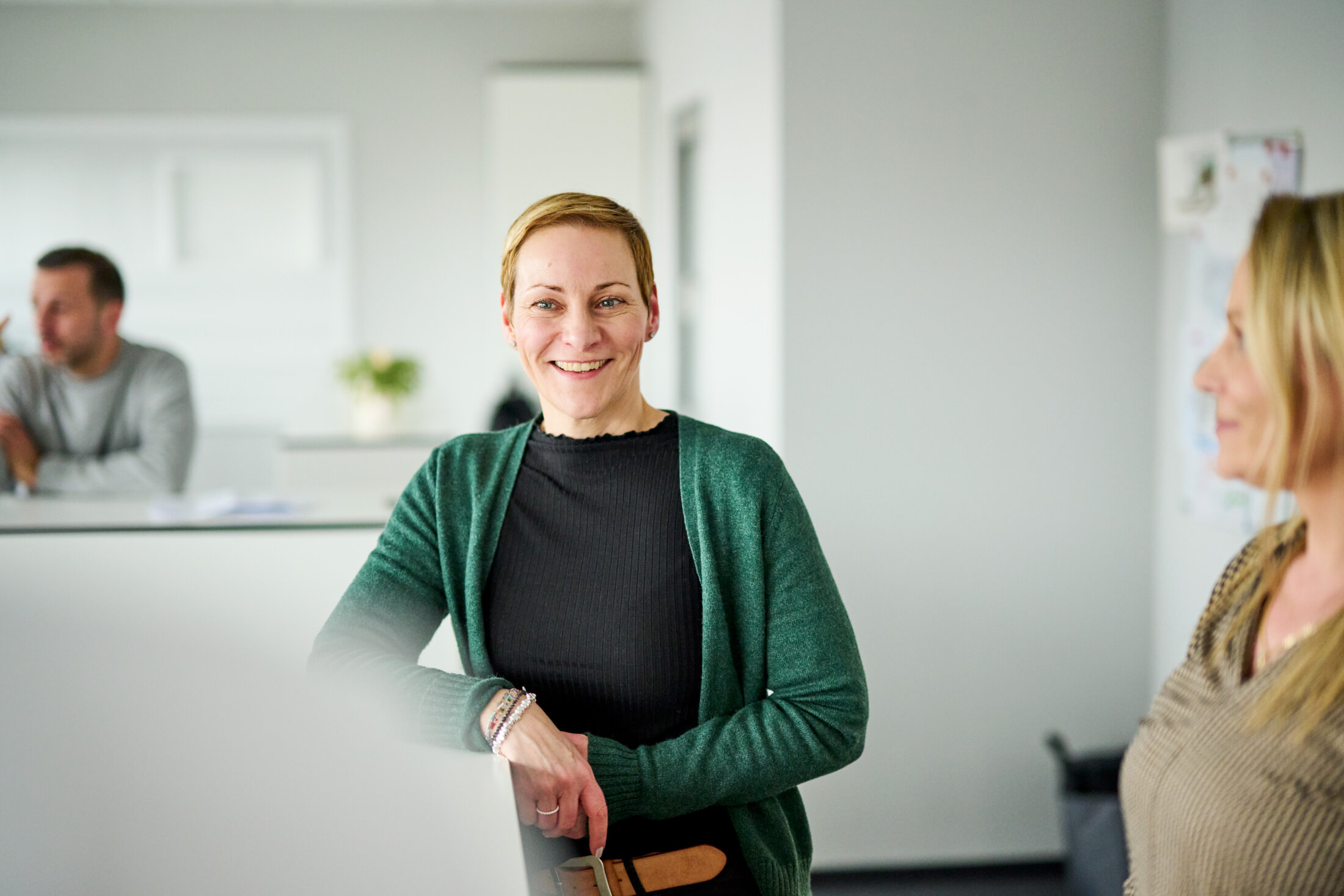 Eine Frau in einem grünen Cardigan lächelt in einem modernen, hellen Büro von HANSETRANS; Im Hintergrund sind zwei weitere Personen zu sehen.