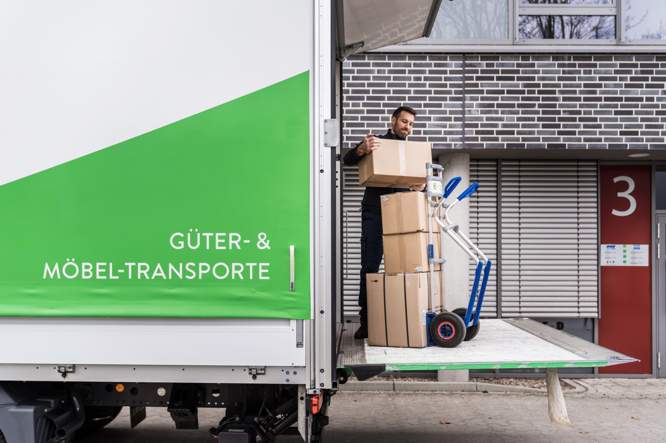 Ein Mann lädt Kartons von einem grün-weißen Umzugswagen mit der Aufschrift „HANSETRANS Güter- & Möbel-Transporte“ auf einen Rollwagen.