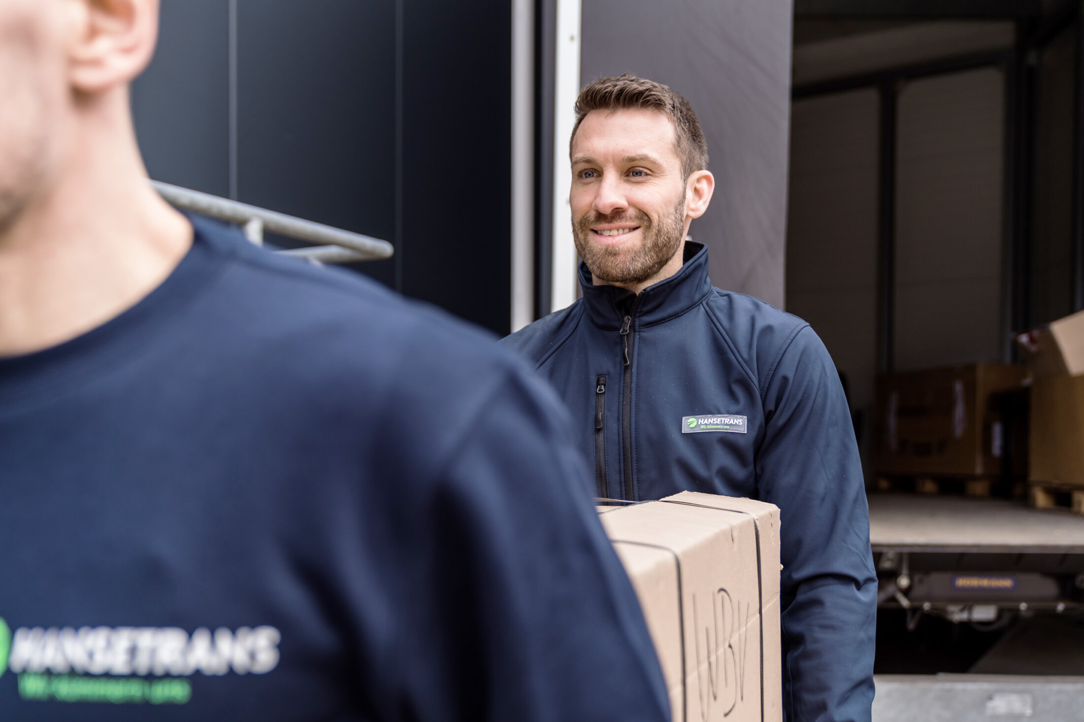 Mann lächelt, während er einen Karton neben einem HANSETRANS-Lastwagen trägt und professionelle Entrümpelung demonstriert; Eine weitere Person steht im Vordergrund und ist bereit, bei der Entrümpelung oder Haushaltsauflösung behilflich zu sein.