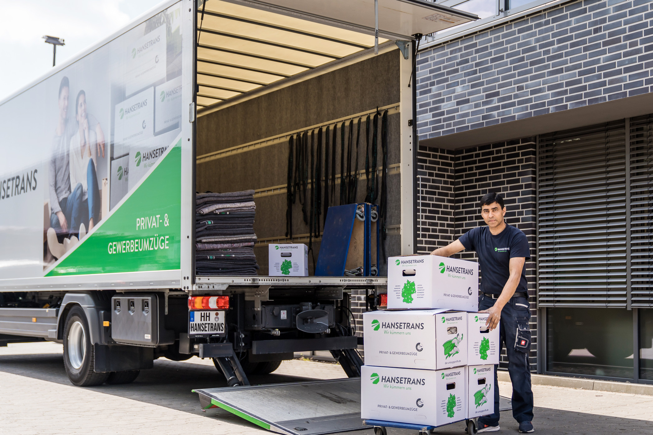 Ein Möbelpacker lädt beschriftete Kartons auf eine Rampe neben einem großen Umzugswagen, der vor einem modernen Gebäude geparkt ist, und sorgt so für einen reibungslosen Seniorenumzug.