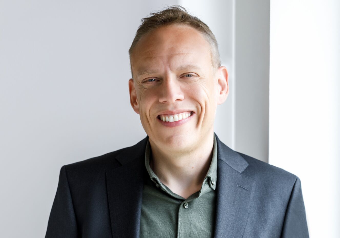 Lächelnde Person in einem dunklen Anzug und grünem Hemd, die neben einer weißen Wand steht.