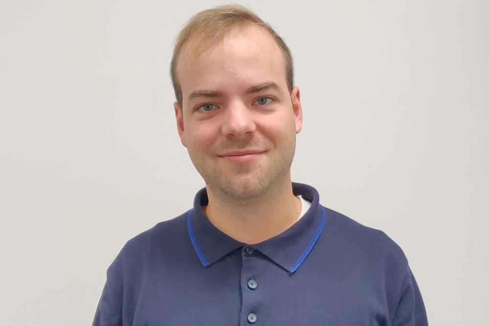 Ein Mann mit kurzen Haaren und Bart trägt ein marineblaues Poloshirt und lächelt in die Kamera. Er steht vor einem schlichten weißen Hintergrund und verkörpert das professionelle und sympathische Image des Frankfurter Kurierdienstes STADTBOTE.