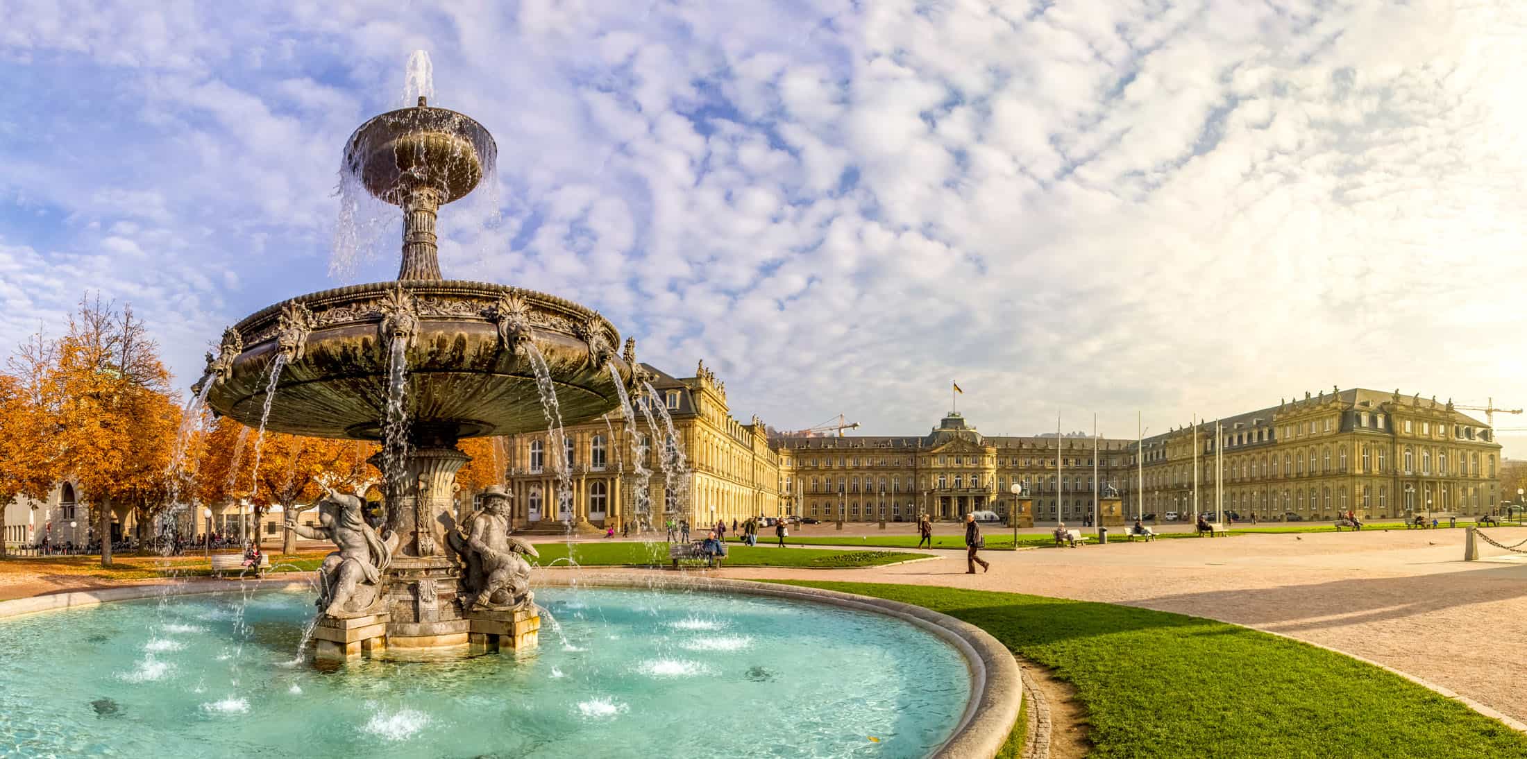 Panoramablick auf einen großen, reich verzierten Brunnen im Vordergrund mit Menschen, die in einem Park spazieren gehen, und einem großen historischen Gebäude unter einem klaren blauen Himmel im Hintergrund.