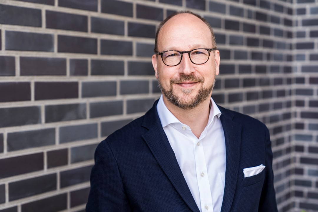 Ein kahlköpfiger Mann mit Brille und Anzug vor einer Ziegelwand.