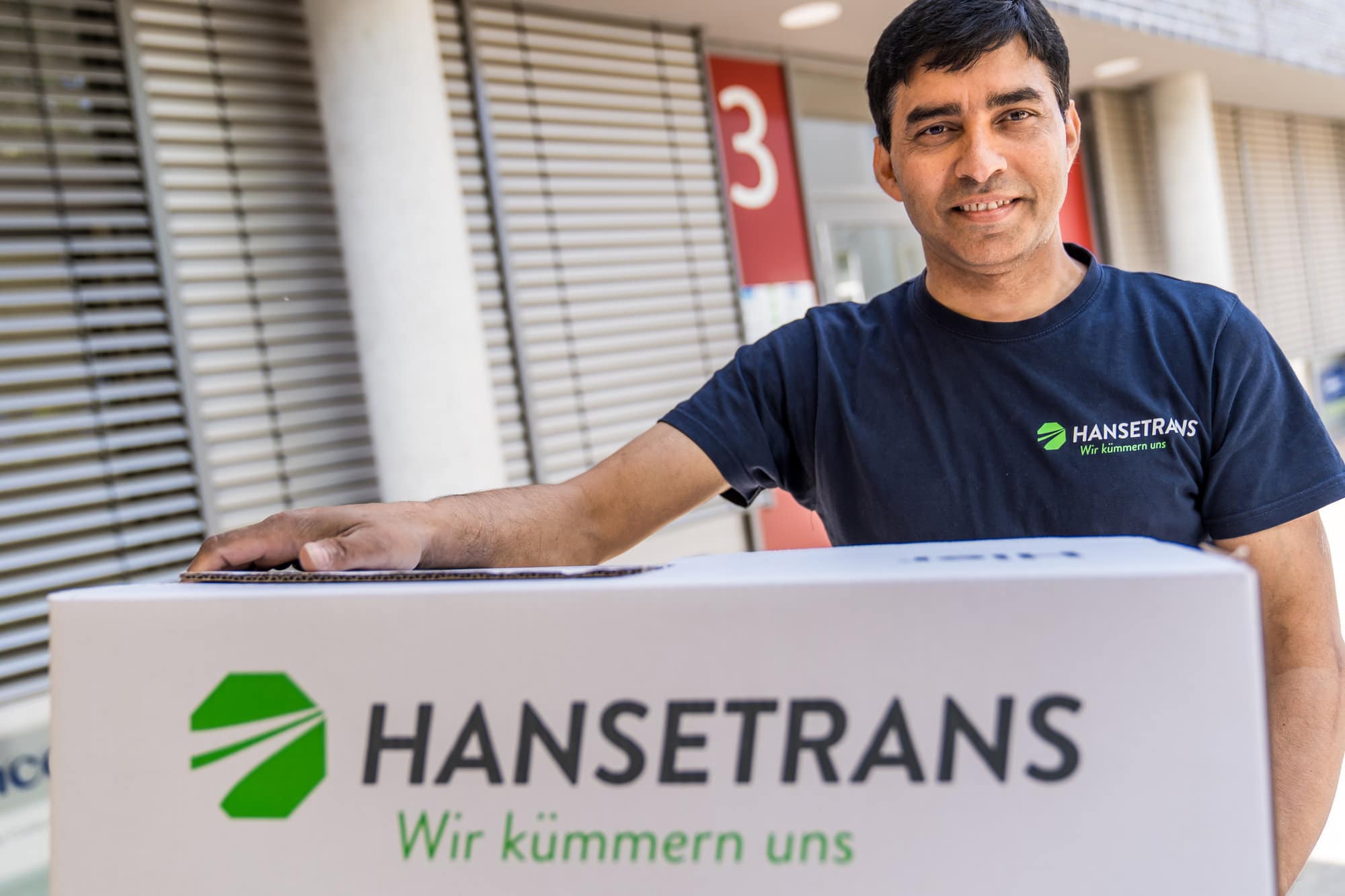 Ein Mann steht neben einer Transportbox mit der Aufschrift „hansetrans“ als Symbol für einen zuverlässigen Transportpartner.